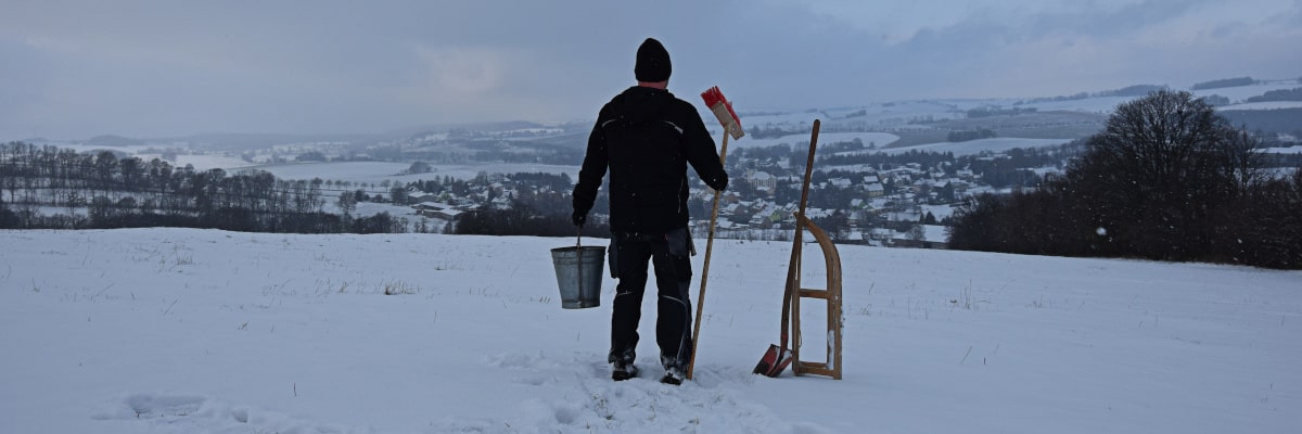 Marek, der Wintermacher – prüfender Blick ins verschneite Großwaltersdorf mit Schlitten, Besen und Eimer im Erzgebirge
