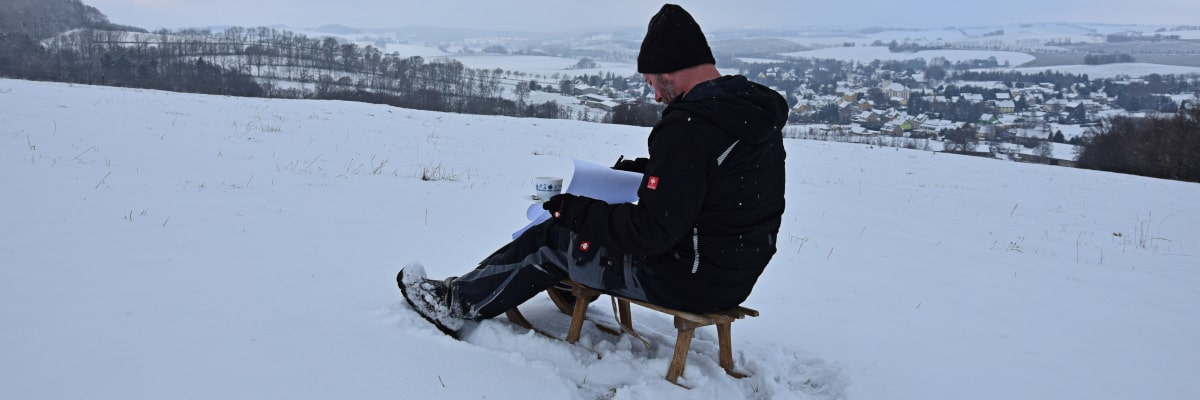 Technikpause im Schnee – Mann genießt Kaffee auf einem Schlitten und blättert entspannt in einem Elektroplan