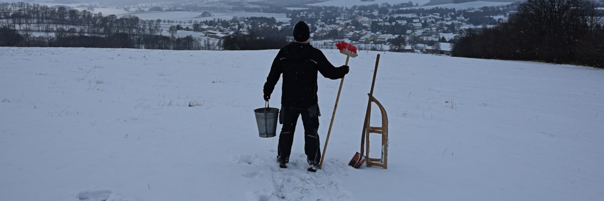 Marek, the maker of winter – man with sledge, broom and bucket stands on a snowy field, gazing watchfully at Großwaltersdorf as if delivering winter